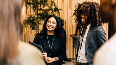 People talk and share ideas in an office setting. Two individuals smile and engage with others while holding papers and wearing lanyards.