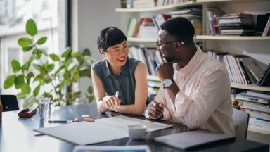 A laughing Japanese businesswoman having a conversation with her African-American coworker while they are sitting at the desk.