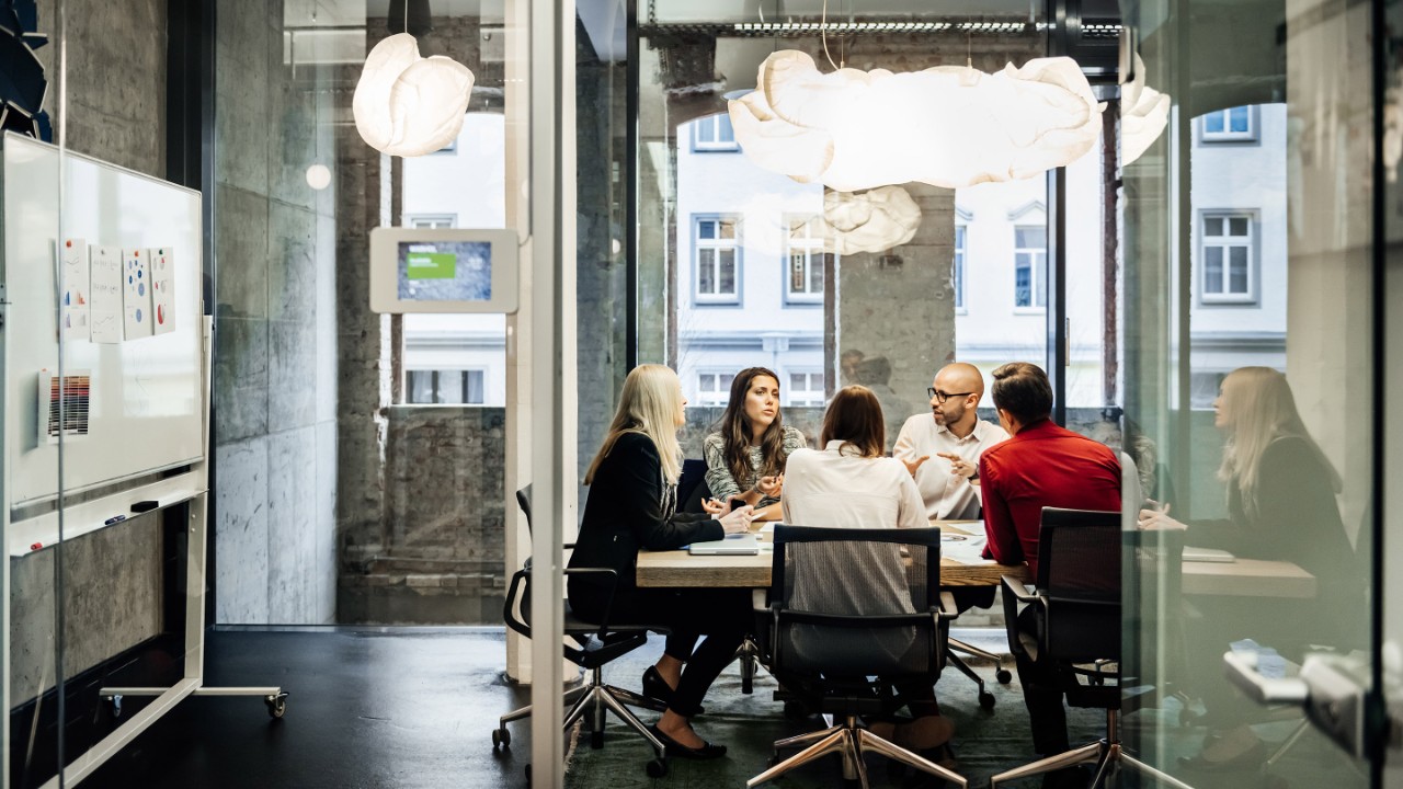 A group of people is sitting at a table during a business meeting in a bright, modern office. The team is talking business while pie charts can be seen hanging on the wall. Big bright windows are seen in the background.