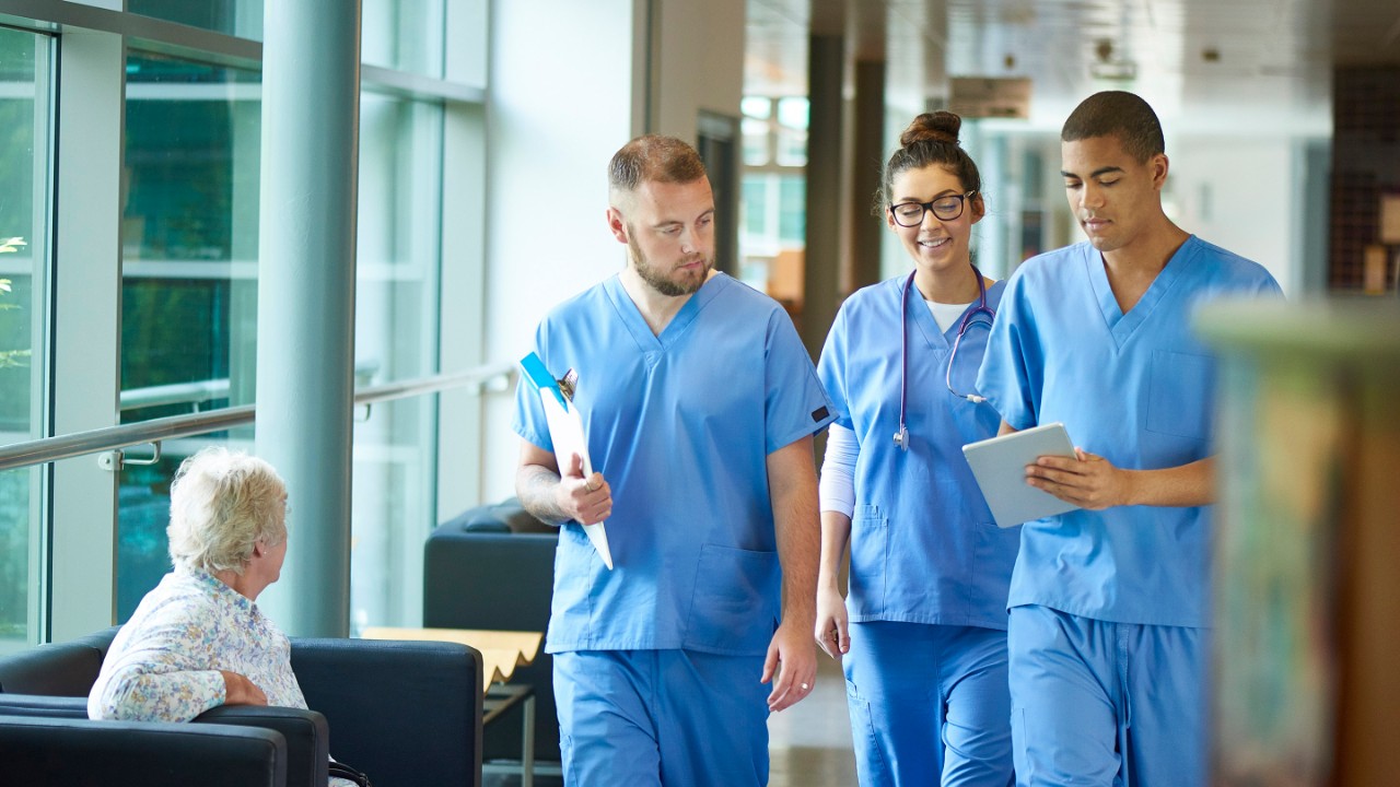three junior doctors walking along a hospital corridor discussing case and wearing scrubs. A patient or visitor is sitting in the corridor as they walk past .