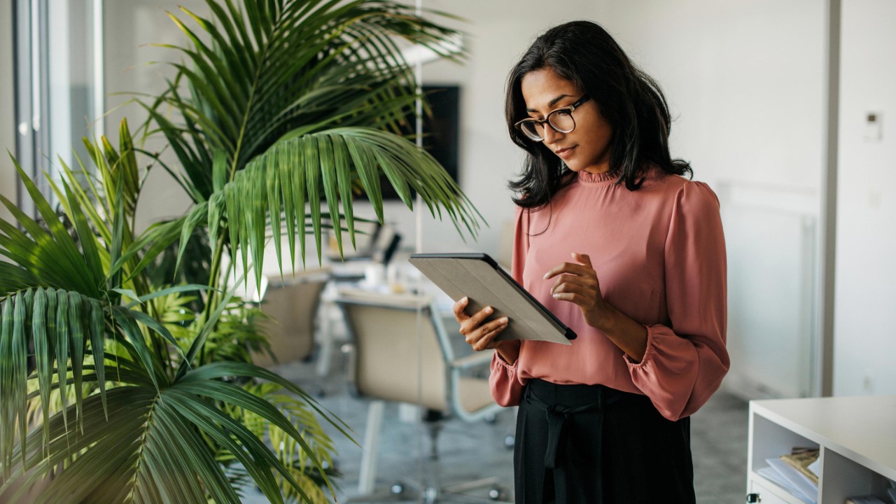 Female executive with medium-length black hair wearing eyeglasses and businesswear standing outside modern meeting room and checking data on tablet.